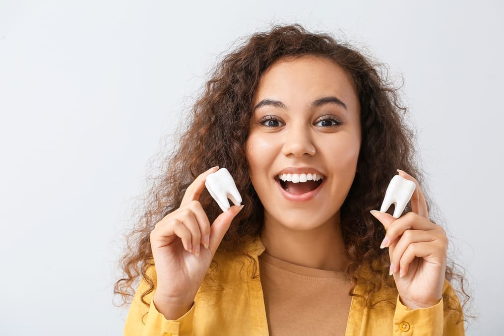 A cheerful young woman with curly hair holds two large tooth models while smiling brightly, symbolizing dental health, education, and the importance of strong teeth – Dentist Columbus Ohio