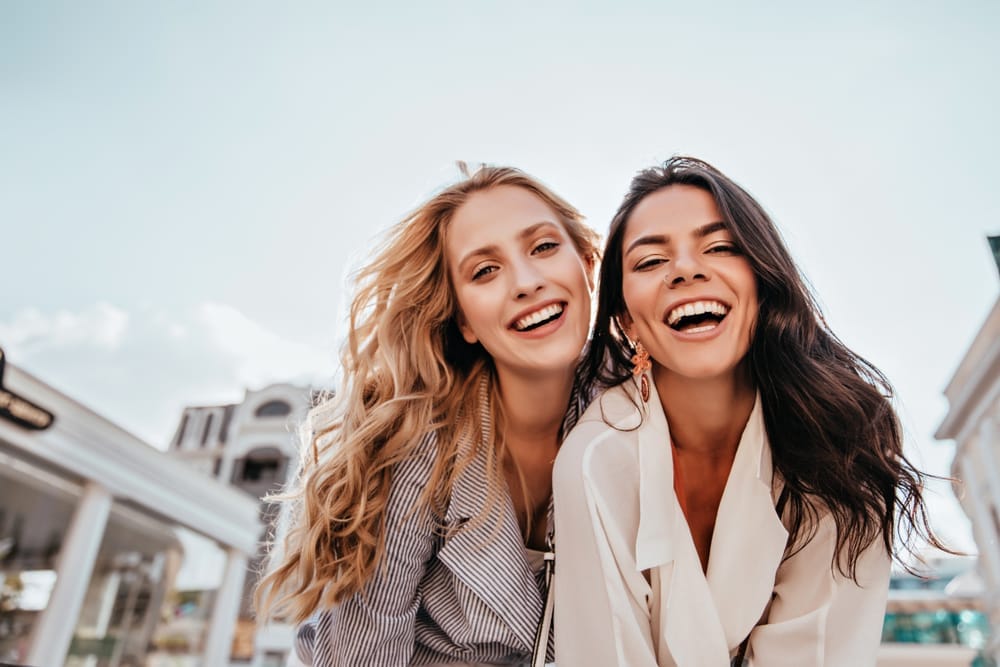 Two cheerful women dressed in fashionable outfits smile brightly while leaning toward the camera outdoors, capturing confidence, beauty, and the joy of a healthy smile – Dentist Columbus Ohio