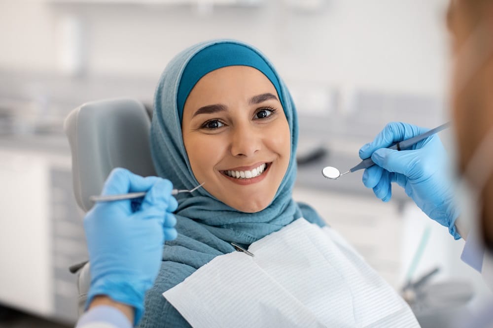 A smiling woman wearing a hijab sitting in a dental chair while the dentist holds tools nearby - Dentist Columbus Ohio​​ 
