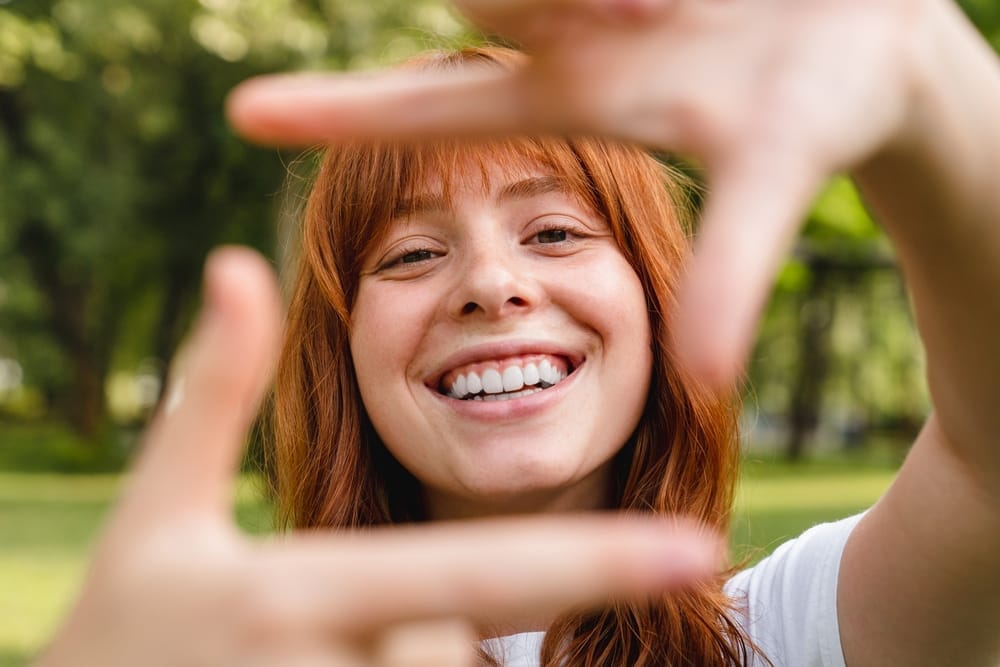 A joyful young woman with red hair smiles widely while playfully framing her face with her hands in a park setting, highlighting happiness, confidence, and the beauty of a healthy smile – Dentist Columbus Ohio