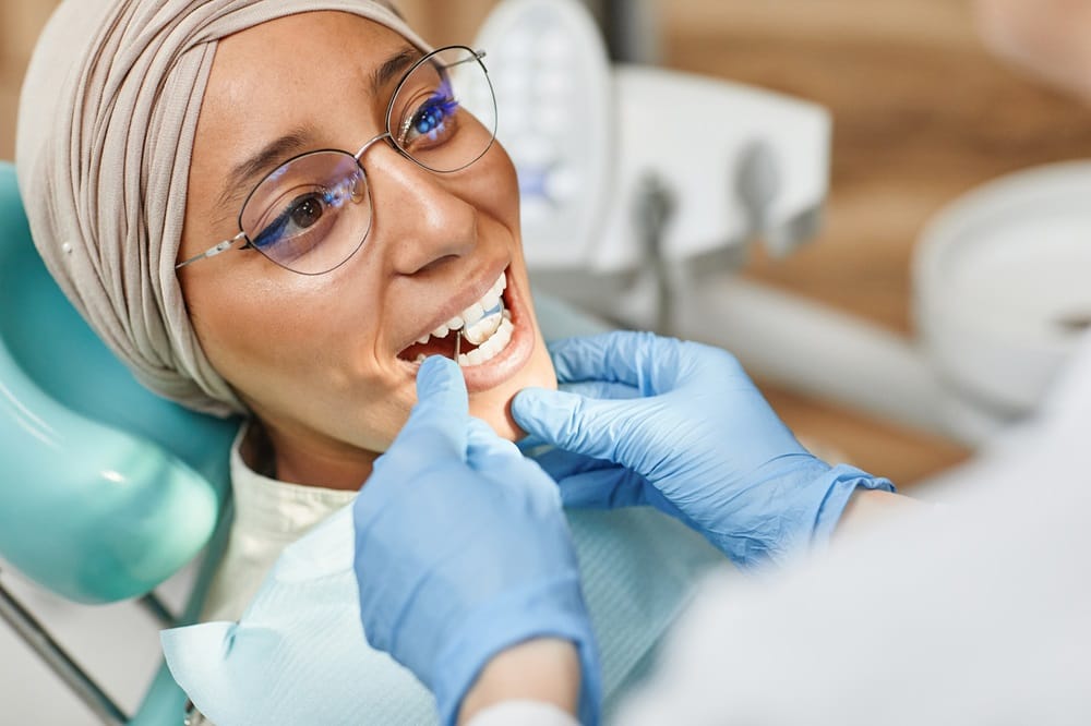 A dentist examines a smiling woman wearing glasses and a headscarf - Dentist Columbus Ohio​​ 